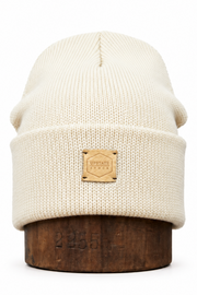 An off-white ribbed knit beanie hat with a tan leather patch on the front cuff, displayed on a dark wooden stand against a white background.