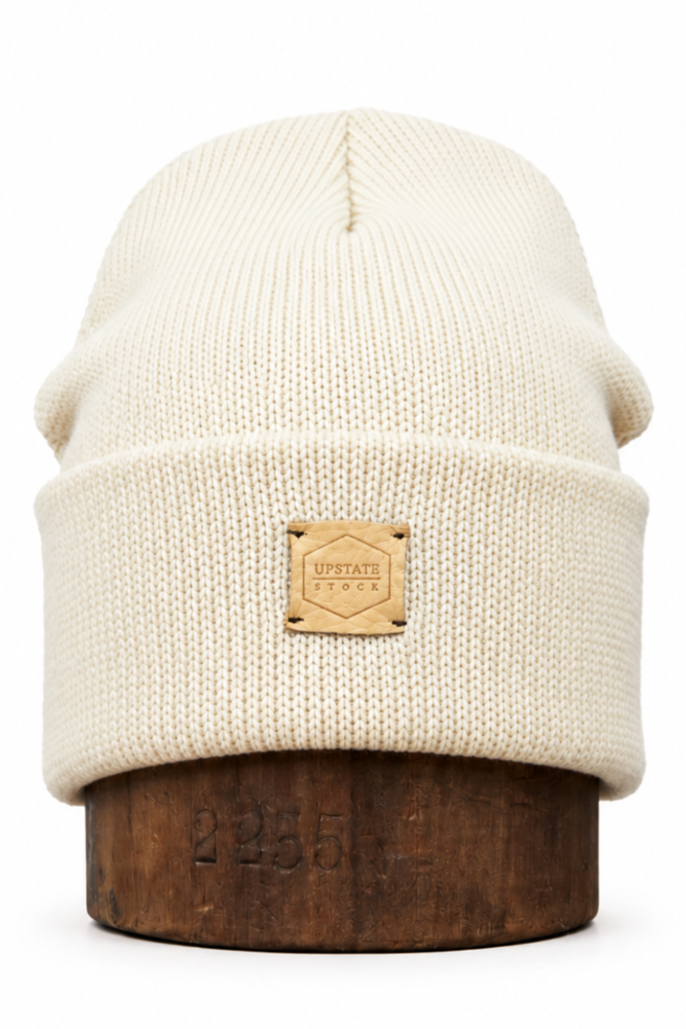 An off-white ribbed knit beanie hat with a tan leather patch on the front cuff, displayed on a dark wooden stand against a white background.