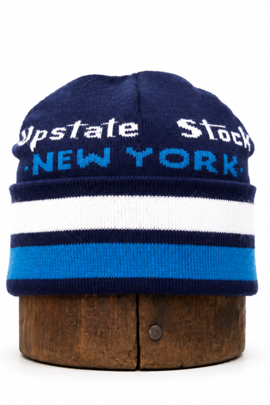 A navy blue knit beanie hat with white and blue stripes, featuring 'Upstate Stock' and 'NEW YORK' text on the front, displayed on a wooden stand against a white background.