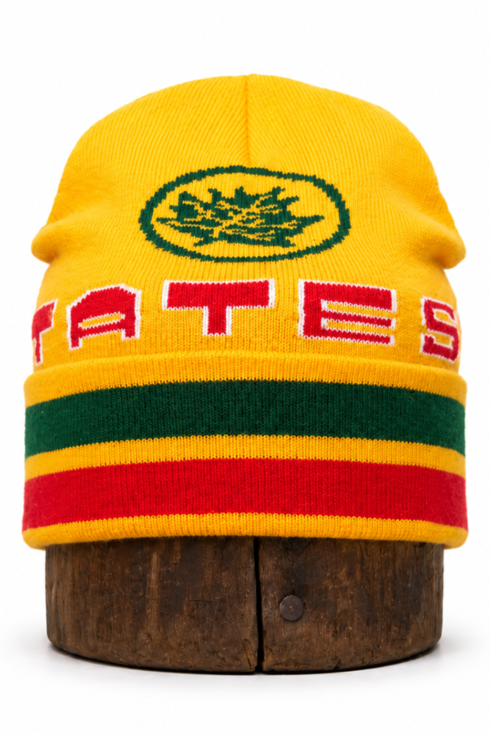 A yellow knit beanie hat with green, red, and yellow stripes, featuring a green emblem and the partial red text 'TATES' on the front, displayed on a wooden stand against a white background.
