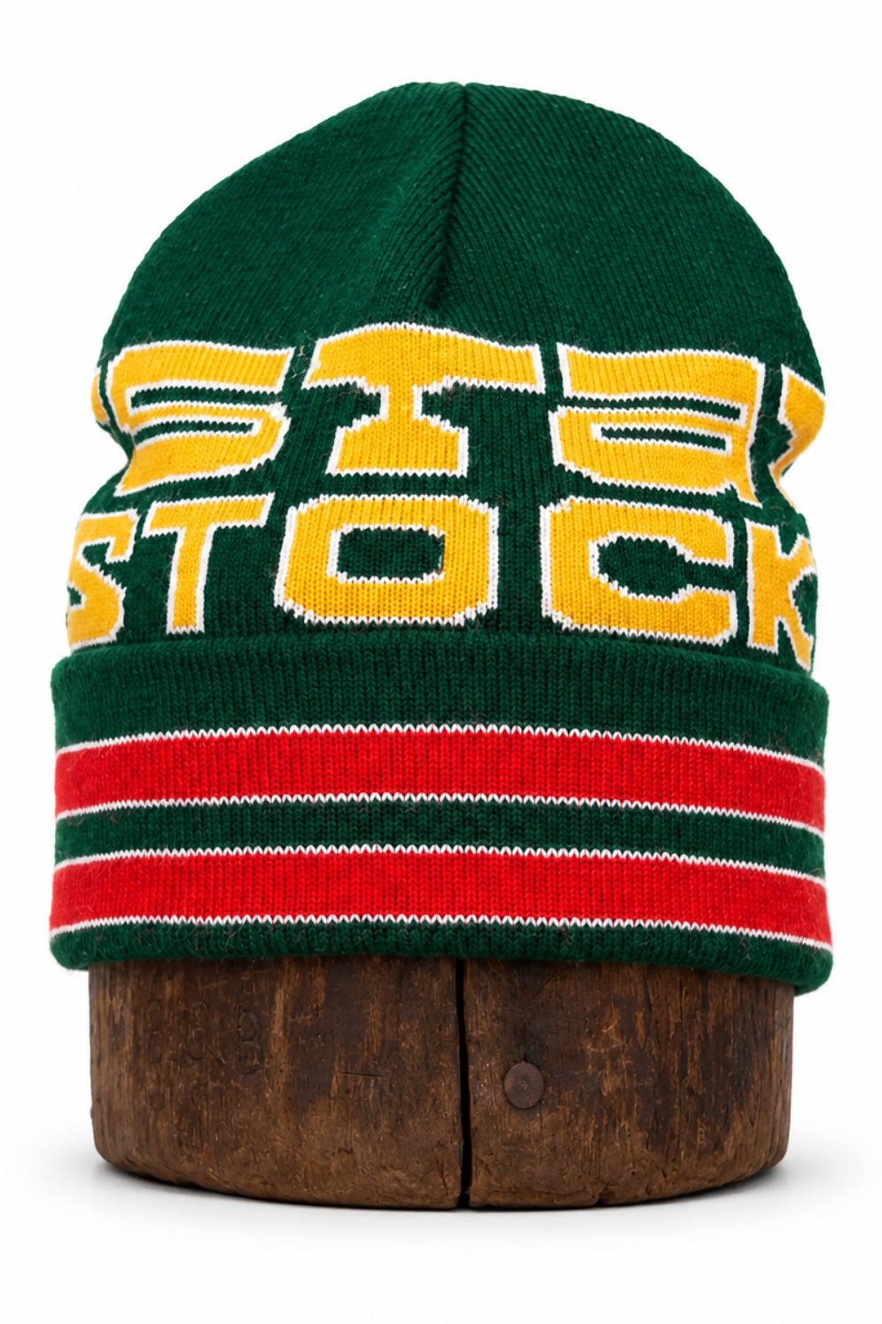 A green knit beanie hat with yellow 'STOCK' lettering and red and white stripes on the brim, displayed on a wooden stand against a white background.