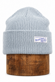 A light blue-gray ribbed knit beanie hat displayed on a cylindrical wooden block against a plain white background. A small white brand patch is visible on the front cuff.