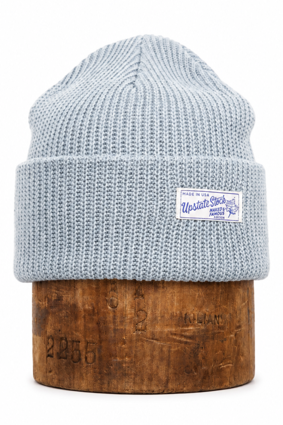 A light blue-gray ribbed knit beanie hat displayed on a cylindrical wooden block against a plain white background. A small white brand patch is visible on the front cuff.