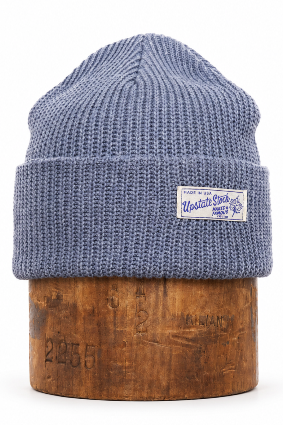 A blue ribbed knit beanie hat with a folded cuff and a small white brand patch, displayed on a wooden stand against a white background.