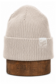 A light beige ribbed knit beanie hat with a folded cuff, displayed on a wooden stand against a plain white background. A small white brand tag is visible on the cuff.