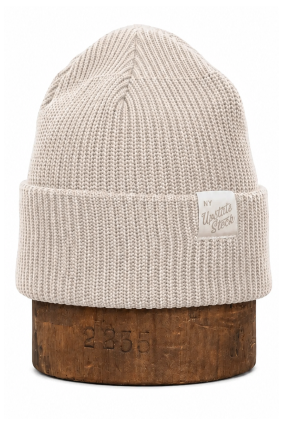 A light beige ribbed knit beanie hat with a folded cuff, displayed on a wooden stand against a plain white background. A small white brand tag is visible on the cuff.