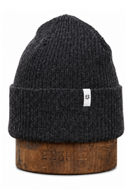 A dark charcoal gray ribbed knit beanie hat with a small white brand tag on the cuff, displayed on a rustic wooden block against a white background.