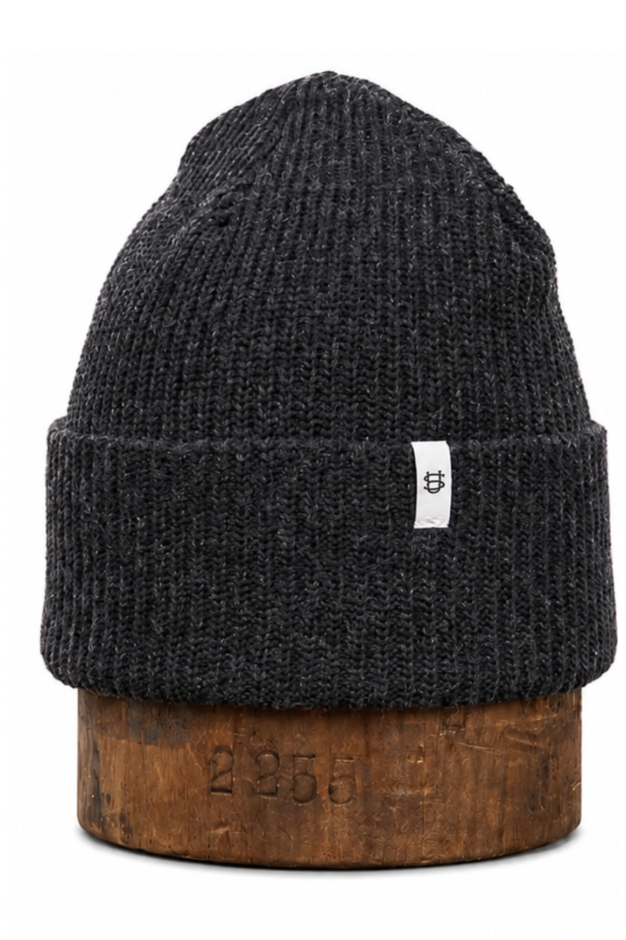 A dark charcoal gray ribbed knit beanie hat with a small white brand tag on the cuff, displayed on a rustic wooden block against a white background.