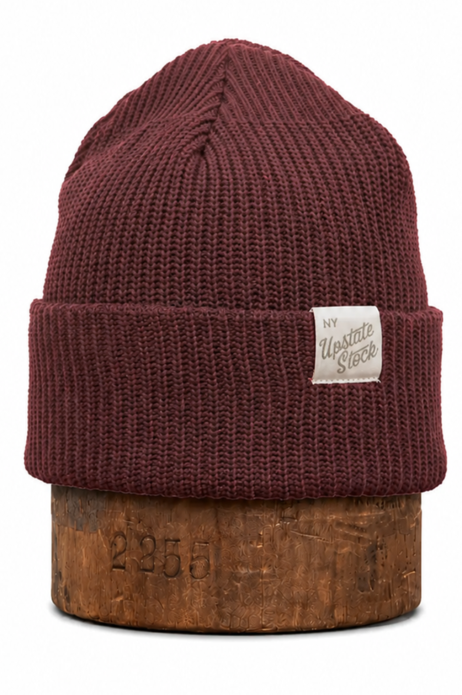 A ribbed knit cuffed beanie hat in a deep burgundy or maroon color, displayed on a wooden block against a plain white background. A small beige fabric brand patch is stitched onto the cuff.