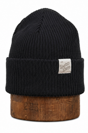 A black ribbed knit beanie hat with a small white 'Western Store' brand patch on the front cuff, displayed on a rustic wooden block against a white background.