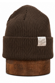 A dark brown ribbed knit beanie hat with a folded cuff, featuring a small white fabric label on the side and displayed on a wooden riser against a white background.
