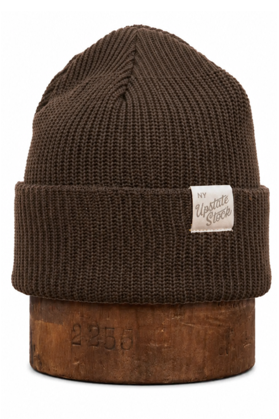 A dark brown ribbed knit beanie hat with a folded cuff, featuring a small white fabric label on the side and displayed on a wooden riser against a white background.