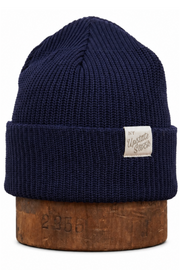 A navy blue ribbed knit beanie hat with a small white rectangular brand patch on the front cuff, displayed on a wooden block against a white background.