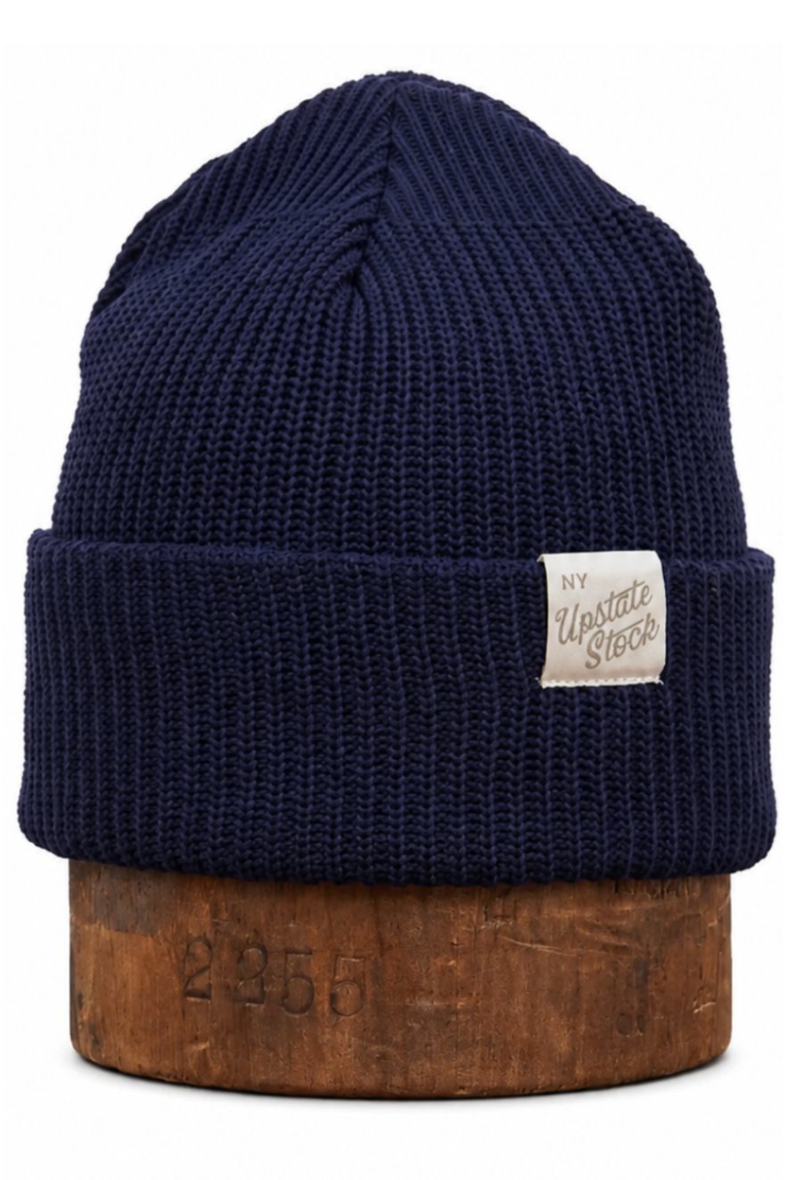 A navy blue ribbed knit beanie hat with a small white rectangular brand patch on the front cuff, displayed on a wooden block against a white background.