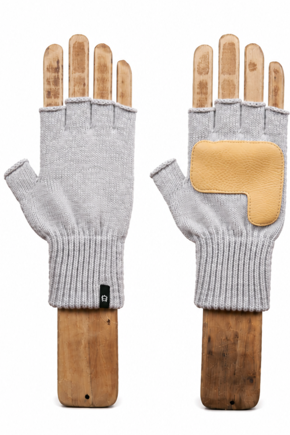 A pair of light gray fingerless knit gloves displayed on wooden hand forms against a plain white background. One glove is shown palm-side with a small black tag, and the other is shown back-side featuring a tan leather patch on the back of the hand.