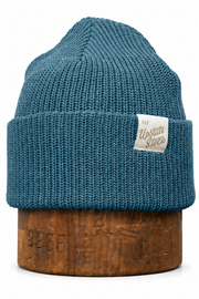 A teal blue ribbed knit beanie hat with a folded cuff, displayed on a wooden stand against a plain white background. A small white woven label on the cuff features the text 'The Misfits Society'.