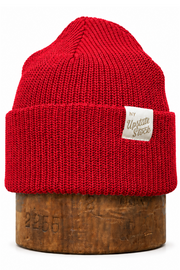 A bright red ribbed knit beanie hat with a small beige fabric label reading 'New United States', displayed on a round wooden stand against a plain white background.