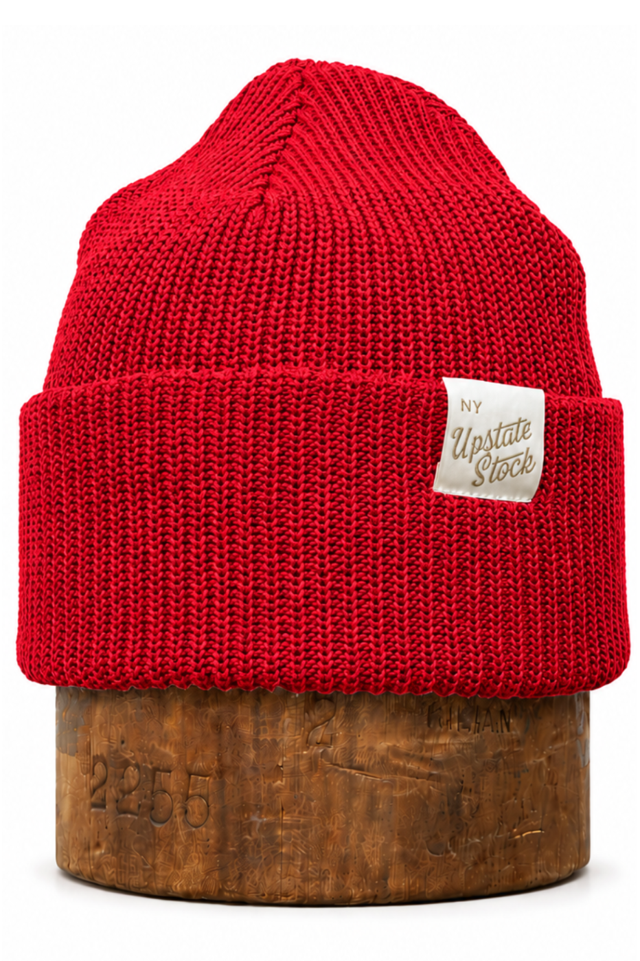 A bright red ribbed knit beanie hat with a small beige fabric label reading 'New United States', displayed on a round wooden stand against a plain white background.