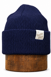 A navy blue ribbed knit beanie hat with a folded cuff, displayed on a wooden stand against a plain white background. A small white fabric label on the cuff reads 'The Upscale Store'.