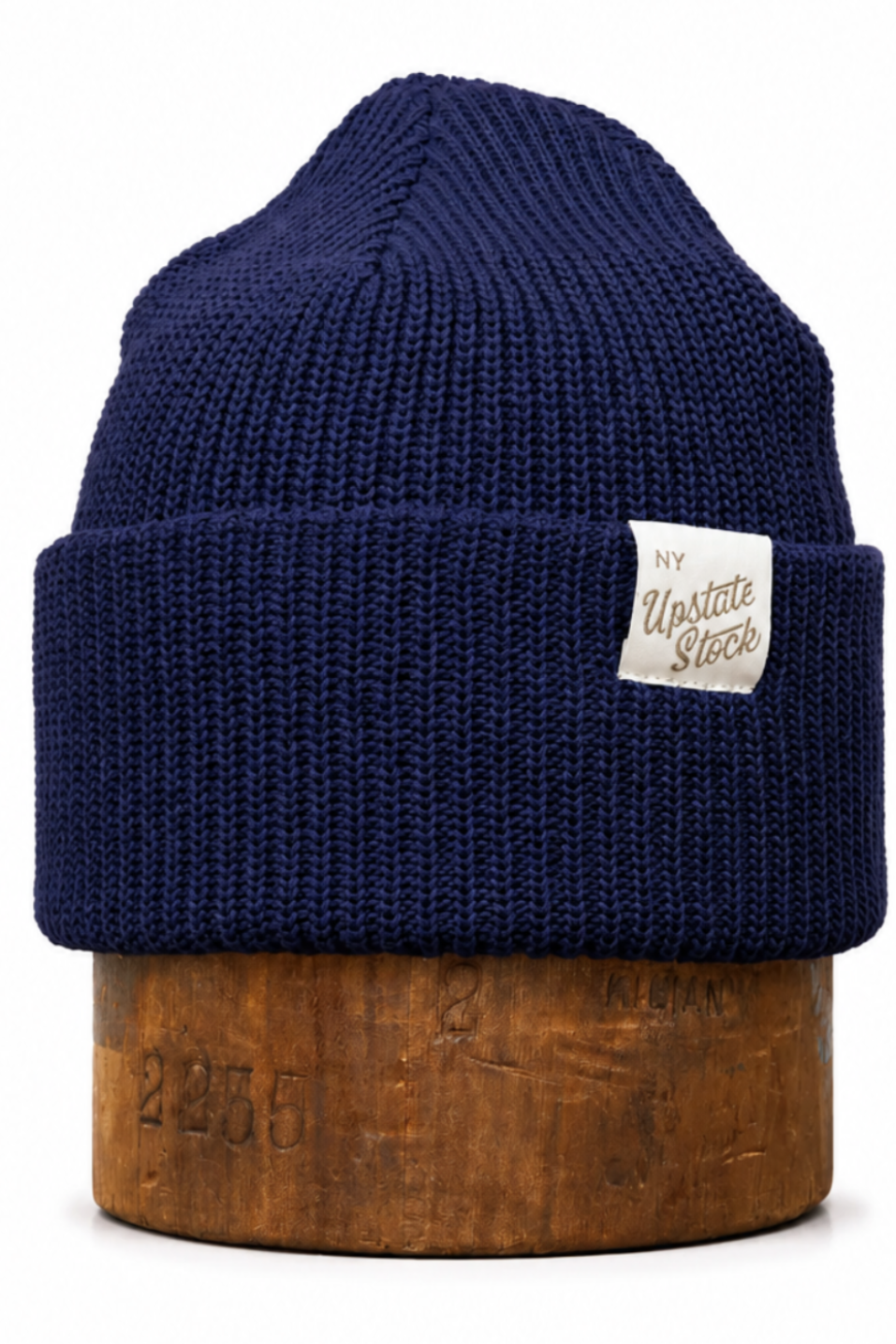 A navy blue ribbed knit beanie hat with a folded cuff, displayed on a wooden stand against a plain white background. A small white fabric label on the cuff reads 'The Upscale Store'.