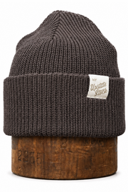 A dark brown ribbed knit beanie hat with a folded cuff, displayed on a wooden stand against a white background. A small white woven label on the cuff reads 'Hustle Sockers'.