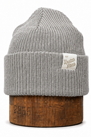 A light gray ribbed knit beanie hat with a folded cuff, displayed on a rustic wooden block against a clean white background. A small cream-colored brand tag reading 'My Update Store' is sewn onto the cuff.