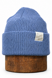 A blue ribbed knit beanie hat with a small white woven label, displayed on a wooden stand against a plain white background.
