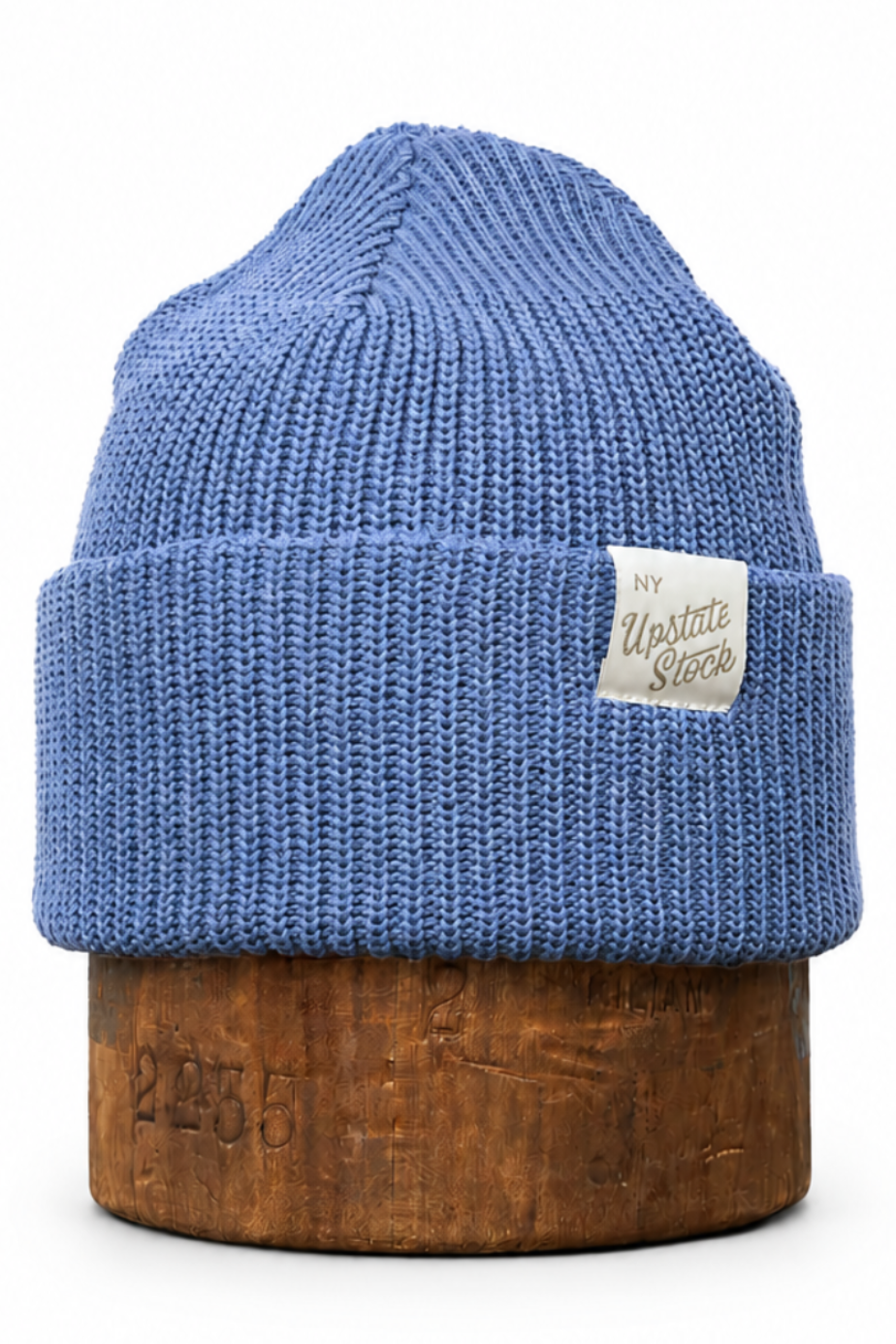 A blue ribbed knit beanie hat with a small white woven label, displayed on a wooden stand against a plain white background.