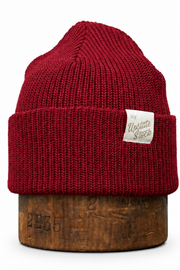 A dark red ribbed knit beanie hat with a small white sewn-on brand patch, displayed on a wooden stand against a plain white background.