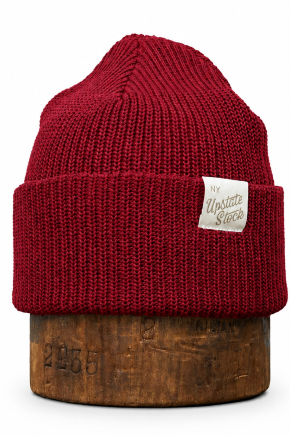 A dark red ribbed knit beanie hat with a small white sewn-on brand patch, displayed on a wooden stand against a plain white background.