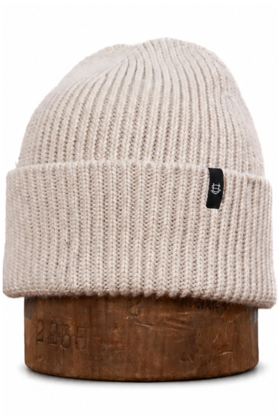 A ribbed knit beanie hat in a light cream or off-white color, displayed on a wooden stand against a plain white background. The beanie features a small black square patch on the side with a white logo.