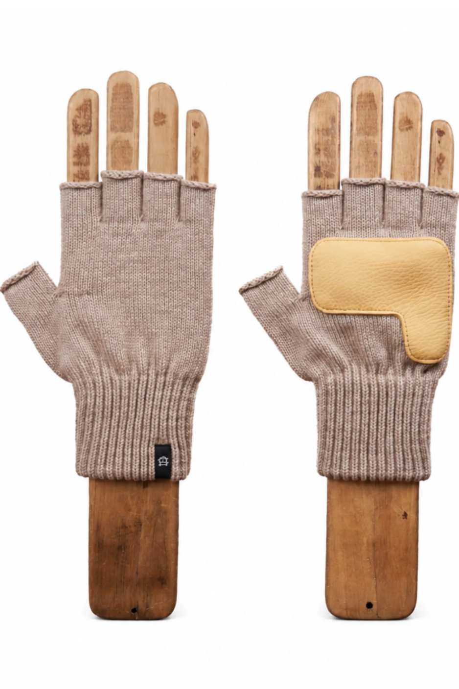 A pair of beige fingerless knit gloves displayed on wooden mannequin hands against a solid white background. One glove is shown palm-side with a small black brand tag, and the other is shown back-side featuring a contrasting light tan leather patch on the back of the hand.