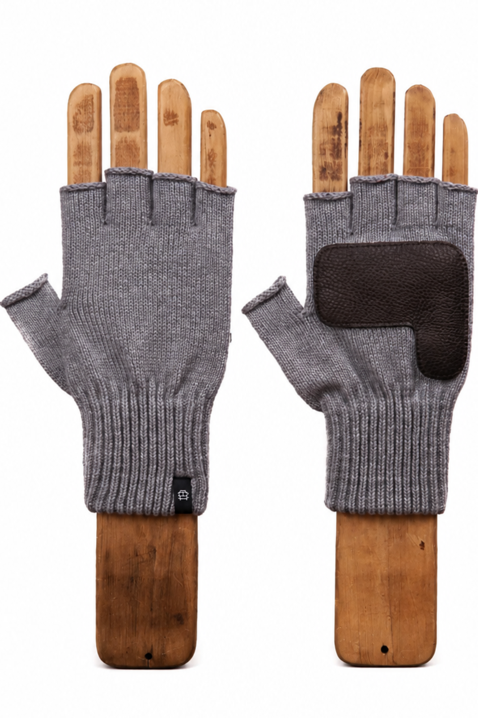 A pair of gray fingerless knit gloves displayed on wooden hand forms against a plain white background. One glove is shown palm-side with a ribbed cuff and a small black tag, and the other is shown back-side with a dark brown reinforced patch on the knuckle area.