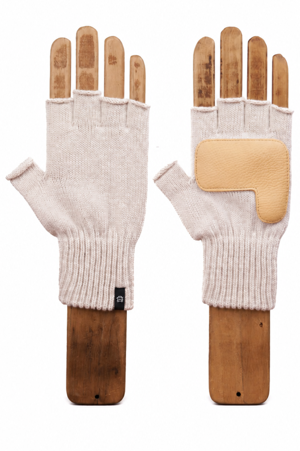 A pair of cream-colored knit fingerless gloves displayed on wooden mannequin hands against a solid white background. One glove is shown from the palm side, featuring a tan leather protective patch on the back of the hand, while the other is shown from the back with the wooden finger forms inserted.