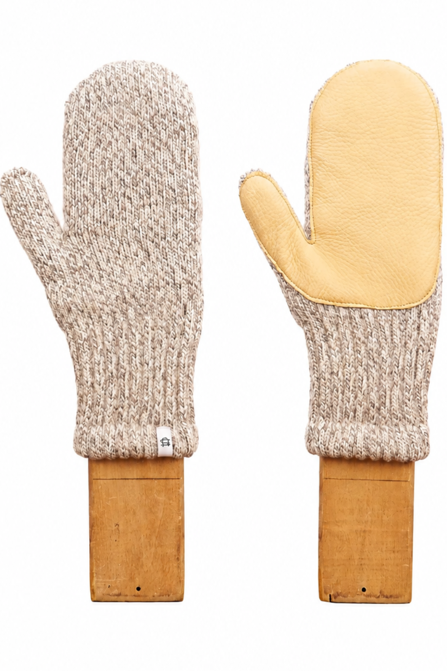 A pair of knit winter mittens displayed side by side on wooden blocks against a plain white background. One mitten is shown from the back, featuring a chunky cream and tan marled knit body, while the other is shown from the front, featuring a smooth, light tan leather palm.