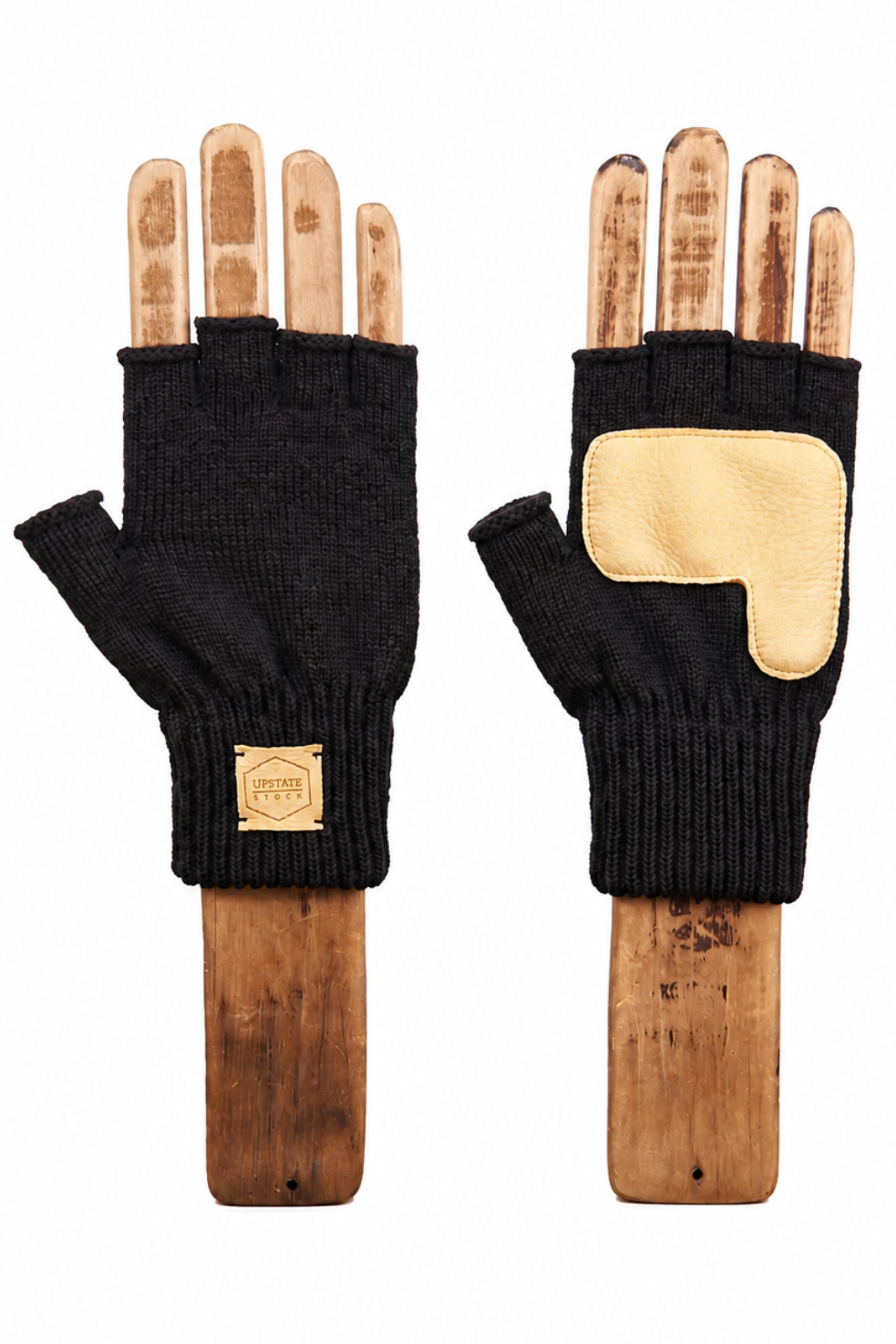 A pair of black fingerless knit gloves displayed on wooden mannequin hands against a plain white background. One glove shows the palm side with a light tan patch, and the other shows the back with a small square brand patch.