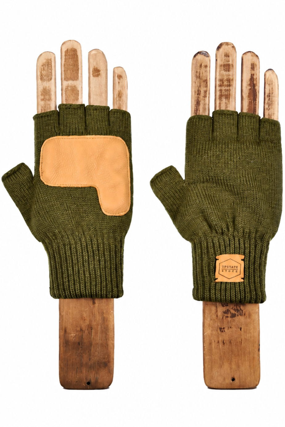 A pair of olive green fingerless knit gloves displayed on wooden mannequin hands against a plain white background. One glove shows a tan leather patch on the back, and the other shows a small tan leather brand patch on the cuff.