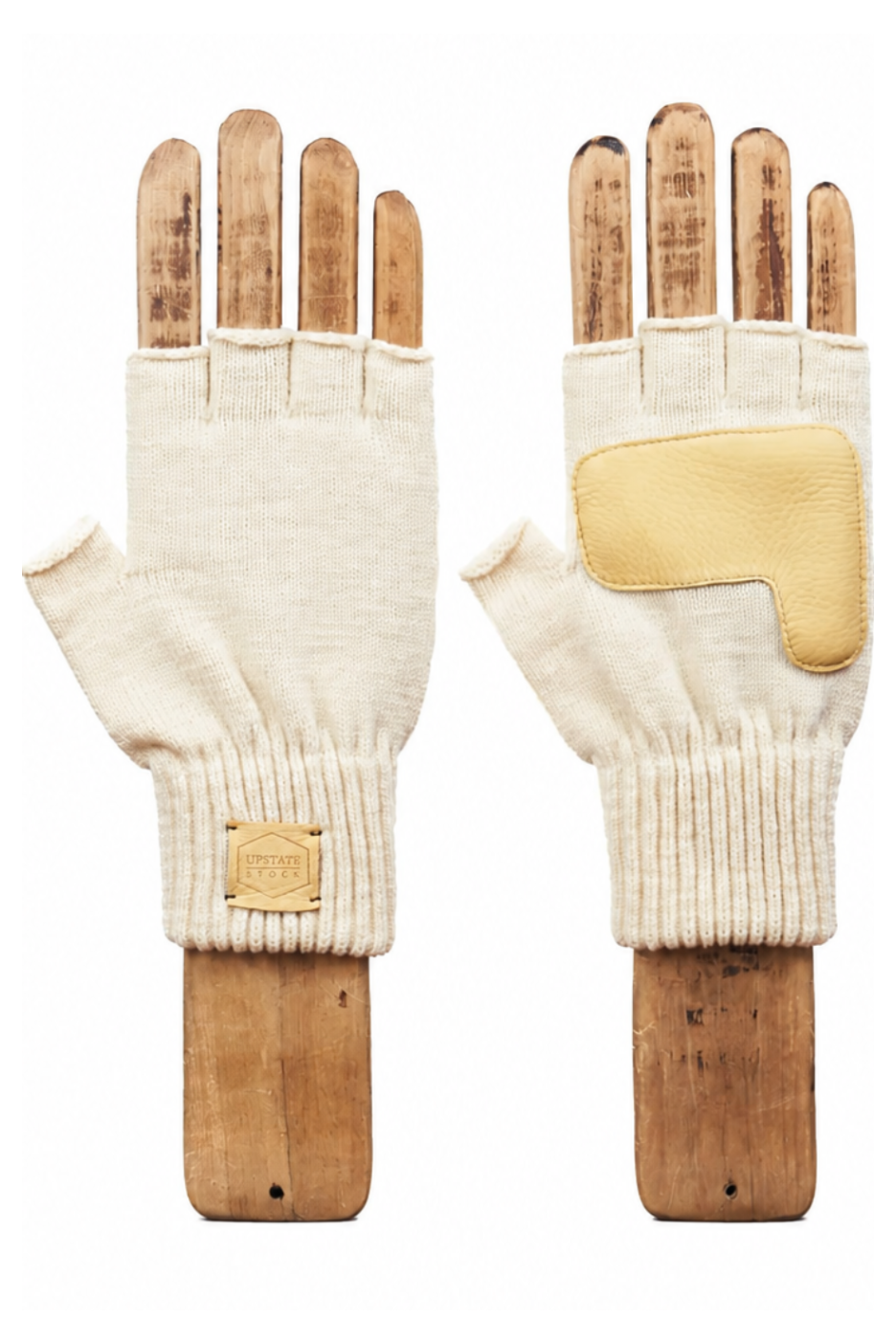 A pair of cream-colored knit fingerless gloves with tan leather patches and a small tan brand patch on the cuff, displayed on wooden hand forms against a white background.