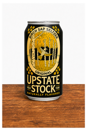 A 16 oz aluminum can of Upstate Stock New York Original Birch Sap Seltzer, featuring a black label with a gold illustration of a birch forest and a figure throwing a pumpkin, set on a wooden surface against a white background.