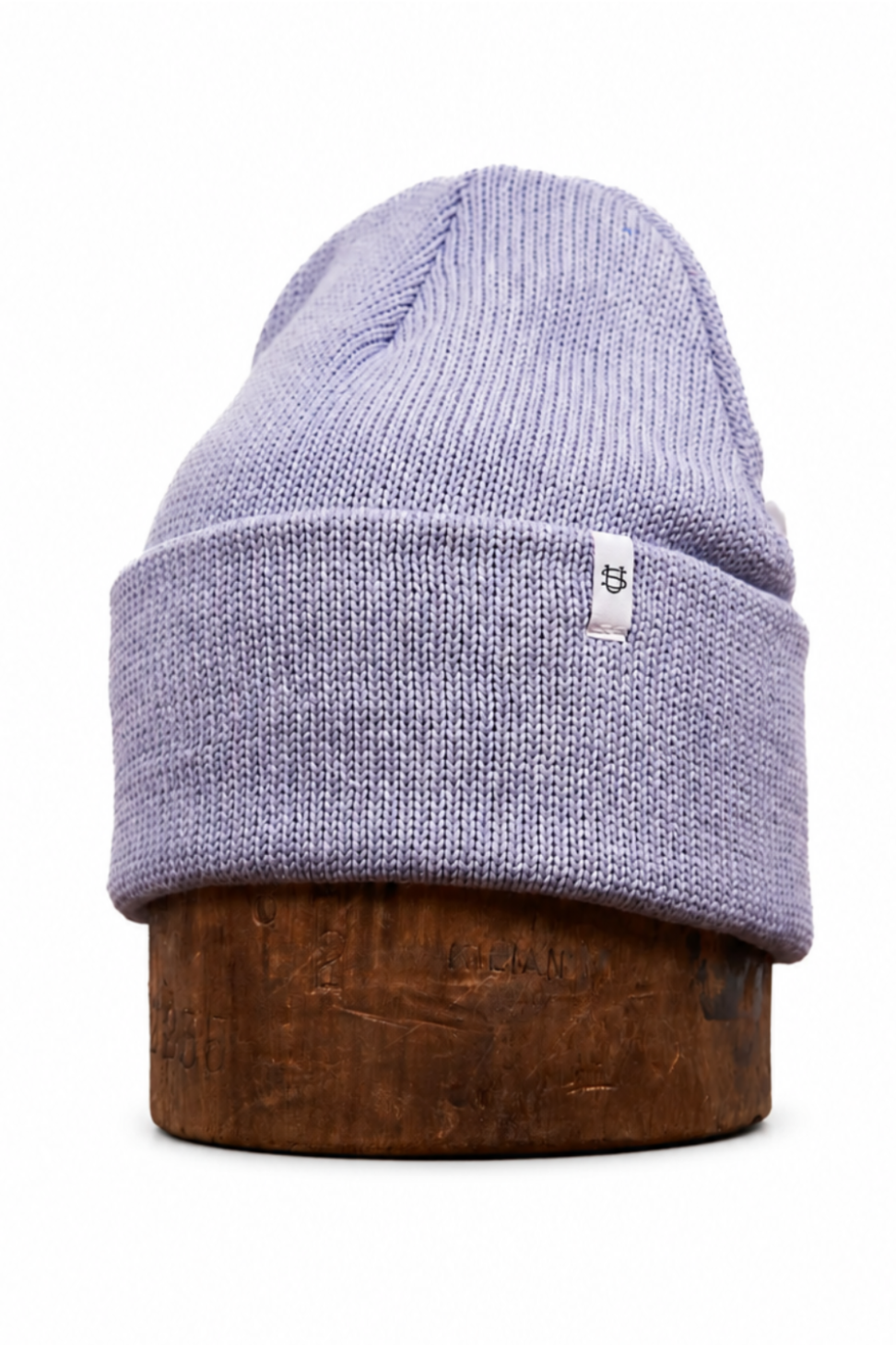 A light lavender knit beanie hat displayed on a round wooden riser against a plain white background.