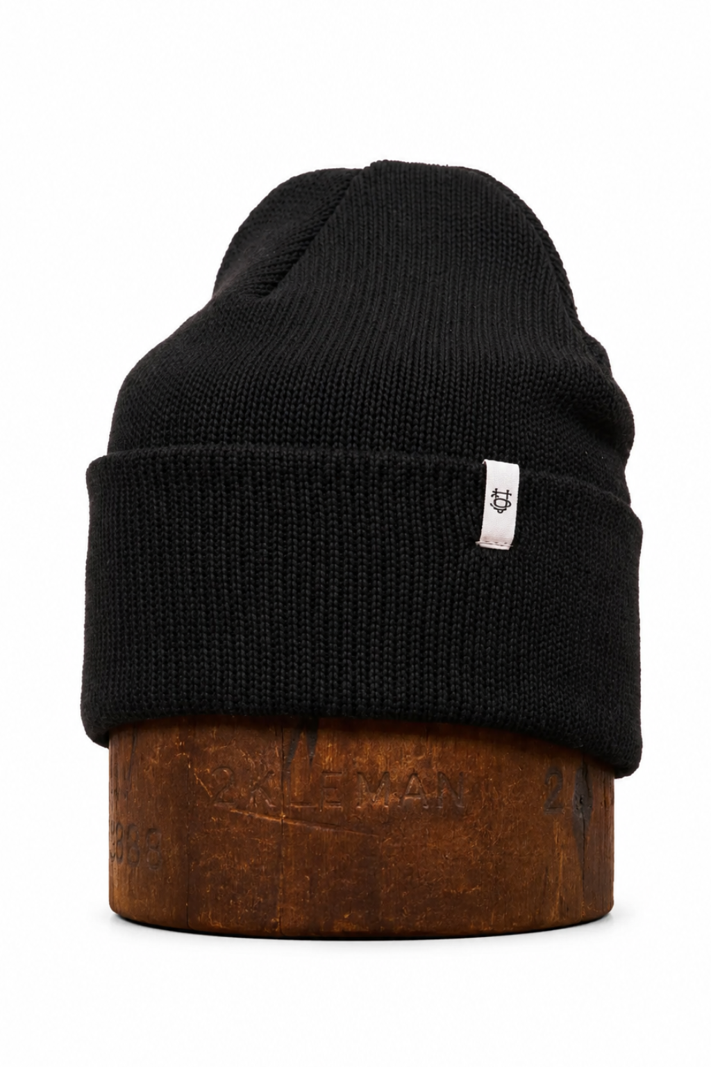 A black ribbed knit beanie hat with a small white rectangular brand tag on the side, displayed on a brown wooden stand against a plain white background.