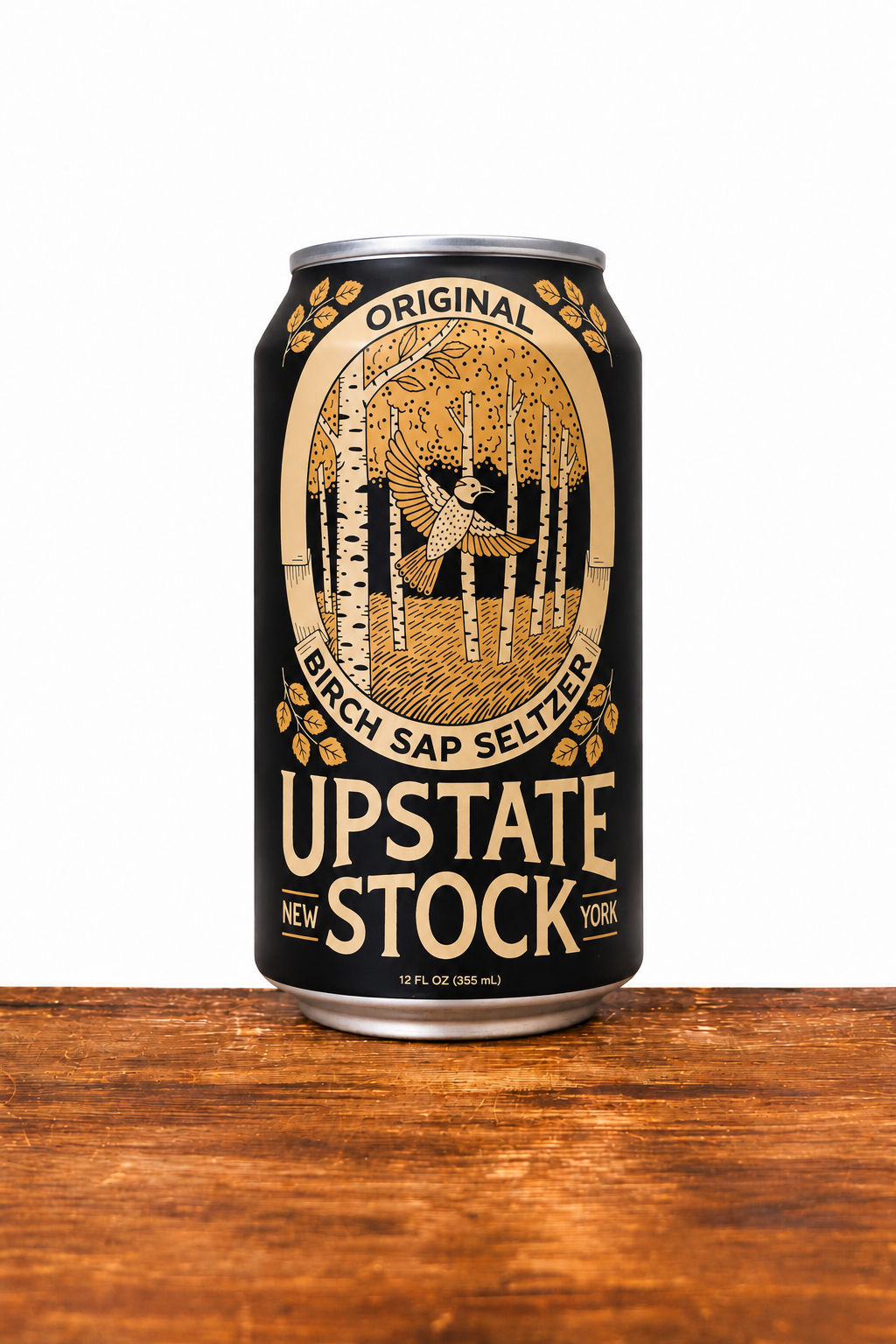 A single can of Upstate Stock Original Birch Sap Seltzer sits on a wooden surface against a plain white background. The can is black with a large tan and gold label featuring a birch tree illustration and the brand name 'Upstate Stock'.