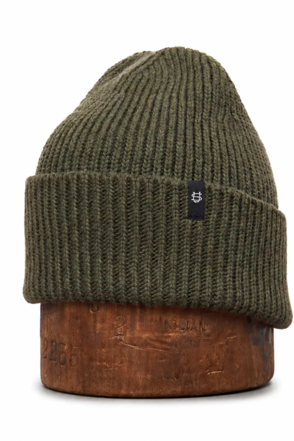 An olive green ribbed knit beanie hat displayed on a round wooden stand against a plain white background.