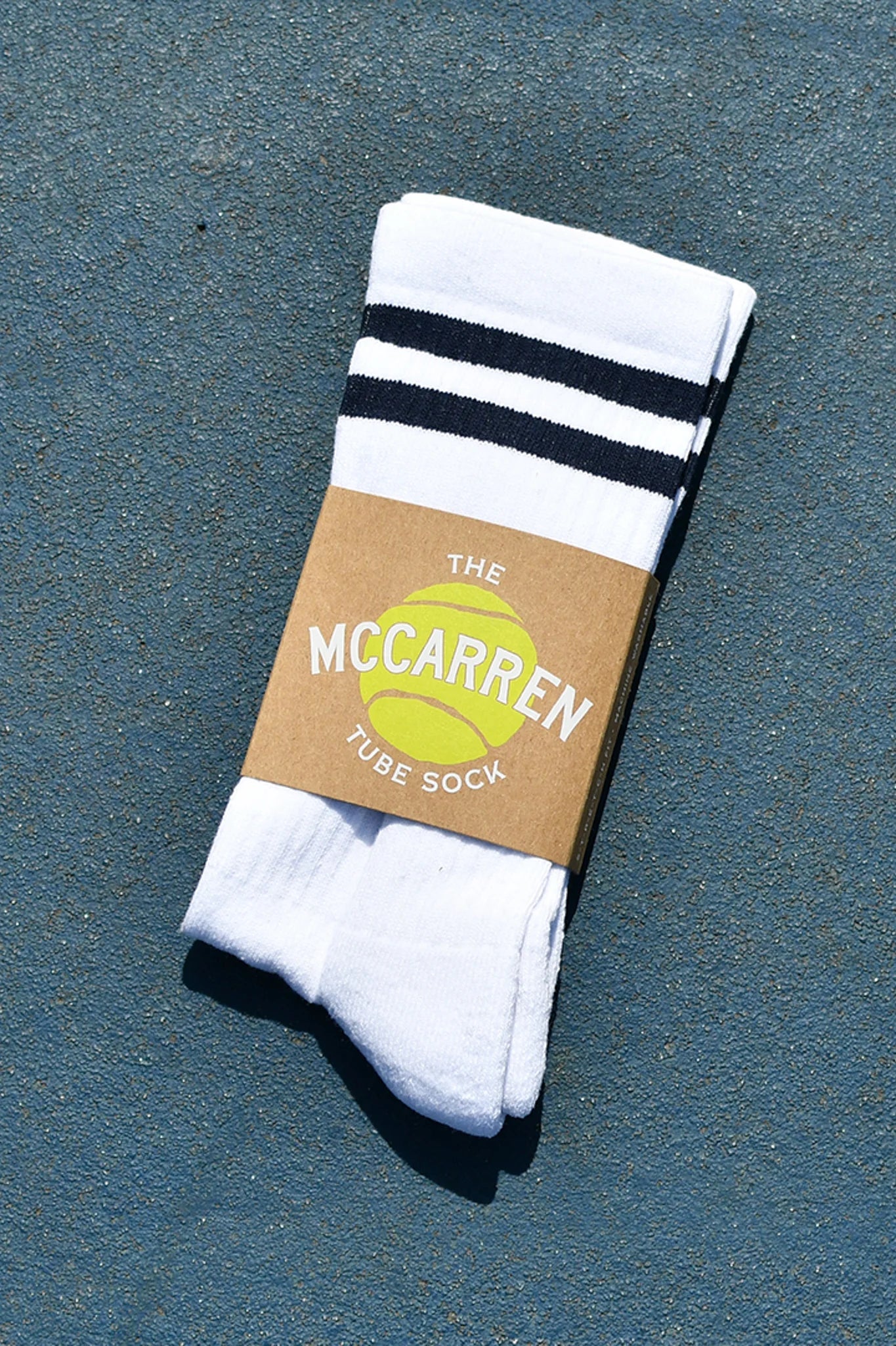 A pair of white tube socks with two black stripes near the cuff, wrapped in a brown paper band that reads 'THE MCCARREN TUBE SOCK' with a yellow tennis ball graphic, placed on a textured blue surface.